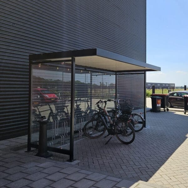 fietsoverkapping bedrijven kopen Fietsoverkapping met glazen zijwanden en een modern metalen dak, geplaatst naast een bedrijfsgebouw. Binnen staan fietsen in een fietsenrek, beschermd tegen weer en wind.