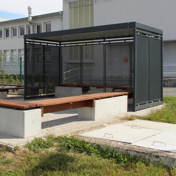 Moderne rookabri met zitgelegenheid direct bestellen Overdekte rookabri met houten zitbanken en glazen wanden naast een bedrijfsgebouw.