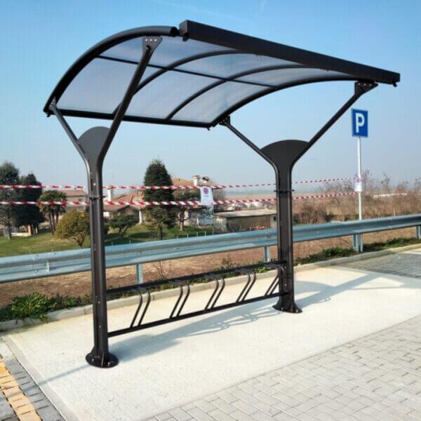 fietsoverkapping-inclusief-fietsenrek-kopen Zijaanzicht van een stalen fietsenoverkapping met gebogen transparant dak en fietsenrek voor 5 fietsen op betonnen ondergrond