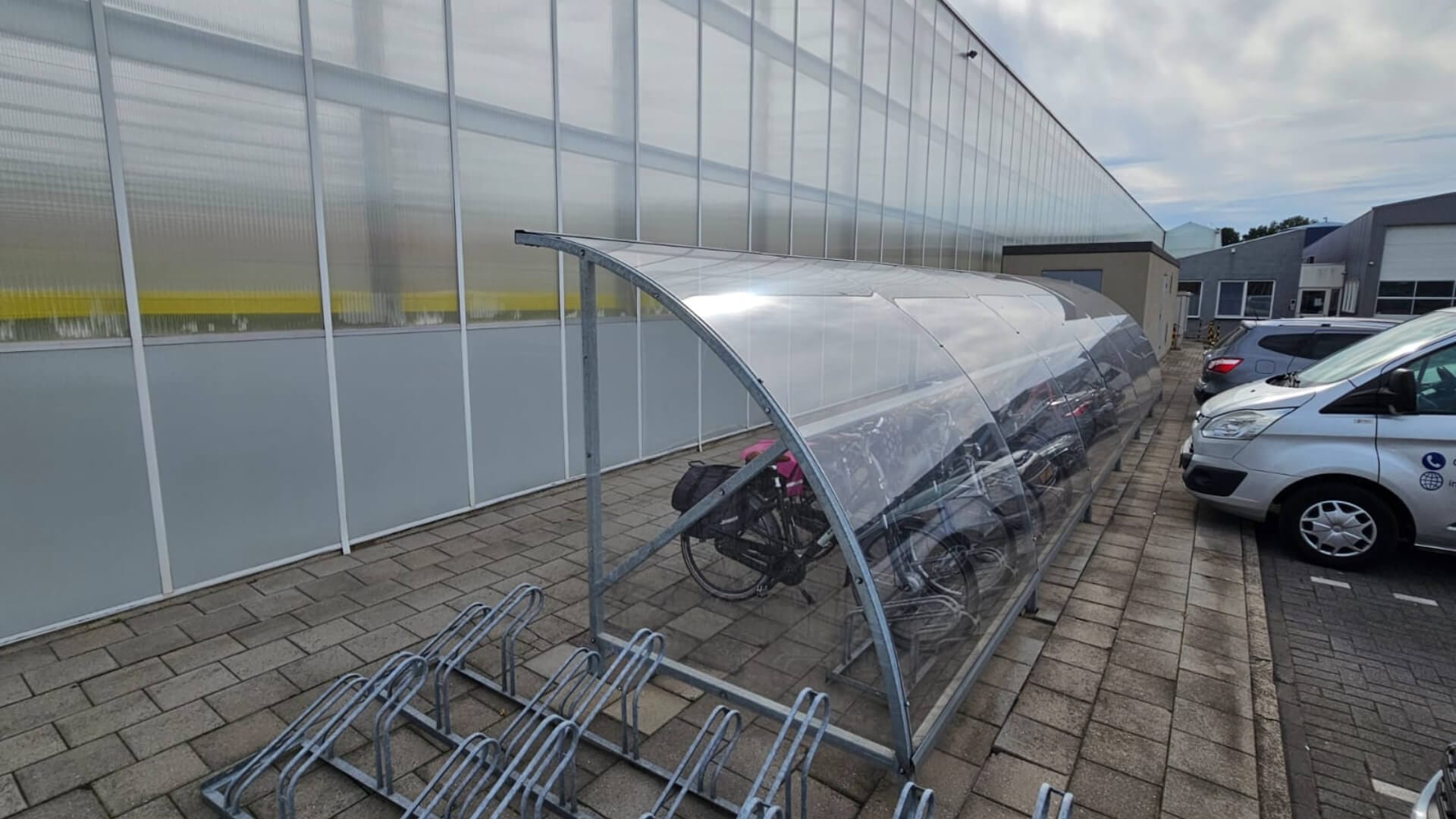 Verzinkte fiets overkapping met helder gebogen dak naast parkeerplaats bij bedrijfsgebouw.