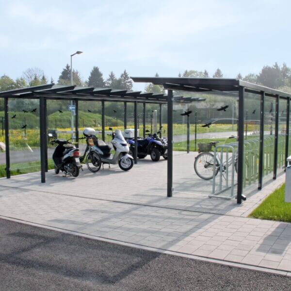 Fietsoverkapping met glazen wanden en universele stallingsmogelijkheden Moderne fietsoverkapping met glazen dak en zijwanden, voorzien van scooters, fietsen en een quad, geplaatst op een nette bestrating.