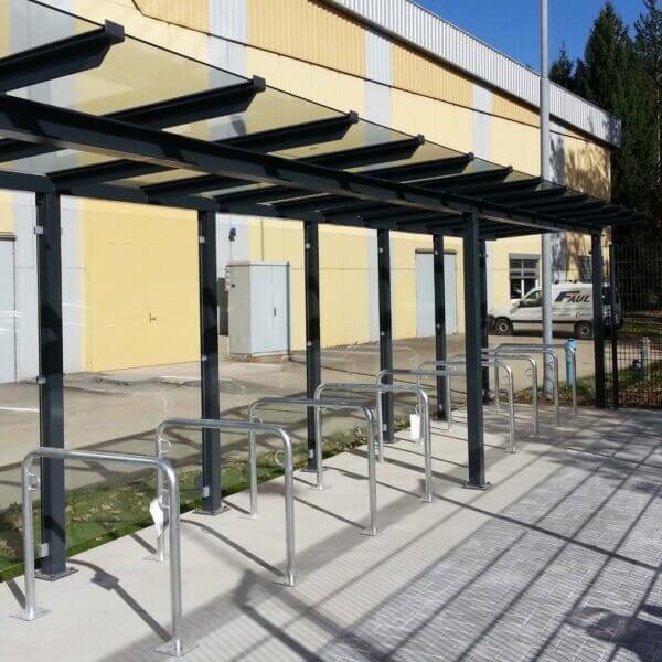 Moderne fietsoverkapping Passau online kopen Fietsoverkapping met glazen dak en geïntegreerde aanleunbeugels, geplaatst op een betonnen ondergrond bij een bedrijfsgebouw.