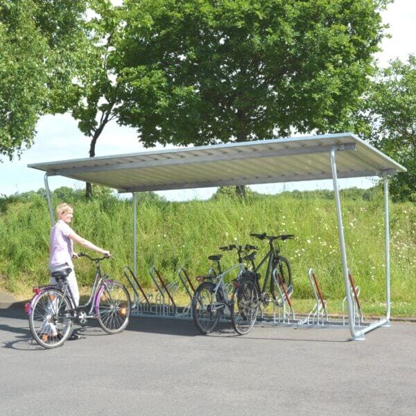 Overdekte fietsoverkapping met metalen dak en verzinkte hoog-laag fietsenrekken, in gebruik door meerdere fietsen, met een vrouw die haar fiets parkeert in een groene buitenomgeving.