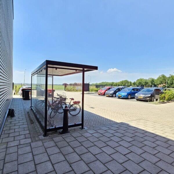 fietsoverkapping voor bedrijven direct online bestellen Glazen fietsoverkapping met metalen frame, geplaatst langs een bedrijfsgebouw. Binnenin staan meerdere fietsen geparkeerd in een fietsenrek. Op de achtergrond is een parkeerplaats met auto's zichtbaar.