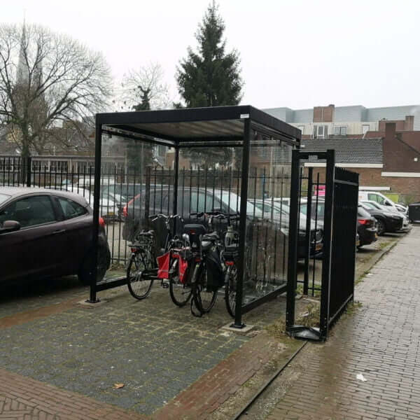 Zwarte fietsoverkapping van staal met ESG glazen wanden, voorzien van optionele fietsrekken en gemonteerd op stevige betonpoeren.