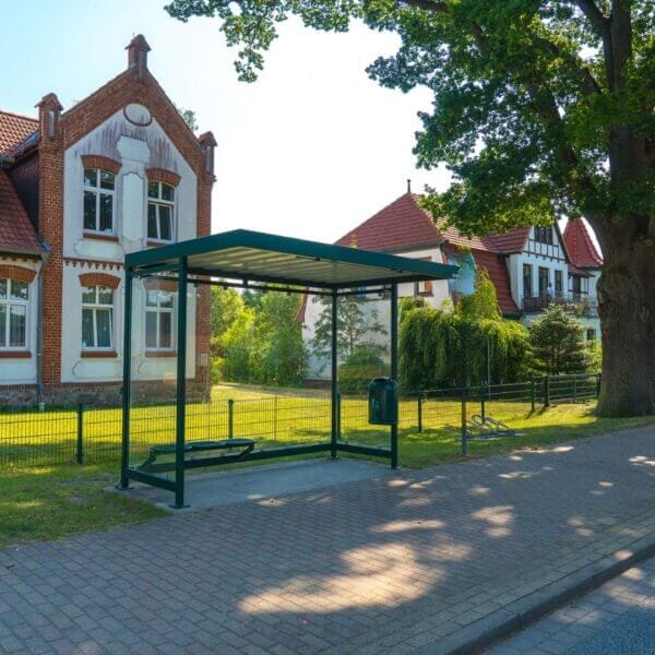 Groen bushokje kopen met glas en stalen constructie Bushokje van staal en glas in RAL 6026, geplaatst in een groene omgeving bij klassieke gebouwen.