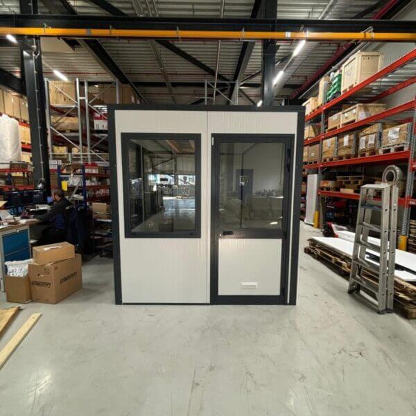 Kantoorunit – Flexibele werkruimte op maat Zelfbouw kantoorunit met raam en deur, geplaatst in een magazijn met stellingen en dozen. De unit heeft witte wandpanelen met zwarte kozijnen en is ontworpen als compacte werkplek.