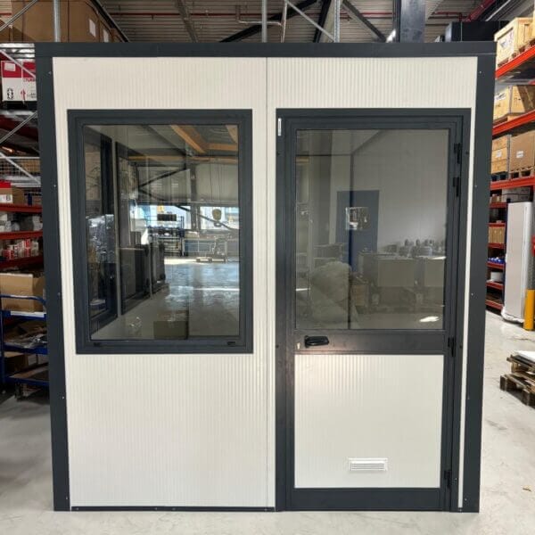 kantoorunit – Snel te plaatsen en goed geïsoleerd Vooraanzicht van een compacte zelfbouw kantoorunit met witte wandpanelen en zwarte kozijnen. De unit heeft een deur met ventilatierooster en een groot raam, geplaatst in een magazijnomgeving.