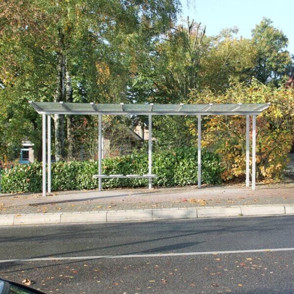 Modern bushokje met glazen dak en stalen frame, geplaatst langs een weg tussen herfstige begroeiing.