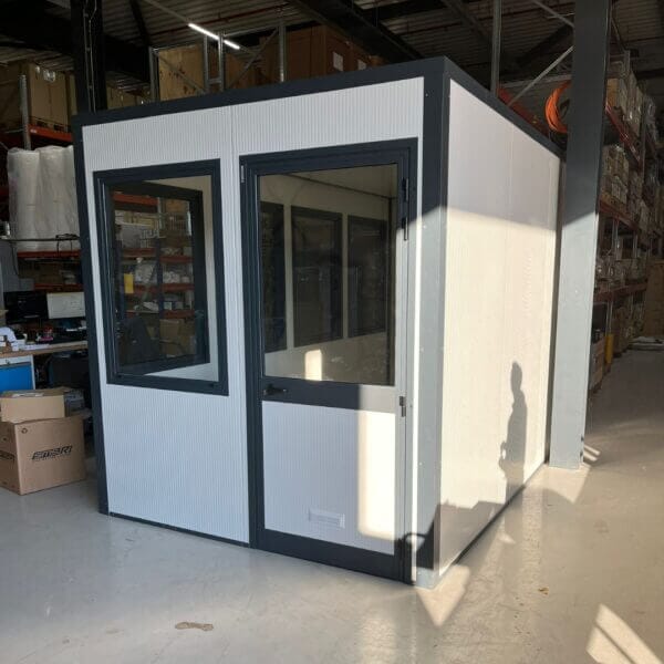 Zelfbouw kantoorunit kopen– Eenvoudig te monteren en isoleren Zelfbouw kantoorunit met witte panelen en donkergrijze kozijnen, geplaatst in een magazijnomgeving. De unit heeft een deur en meerdere ramen voor lichtinval.