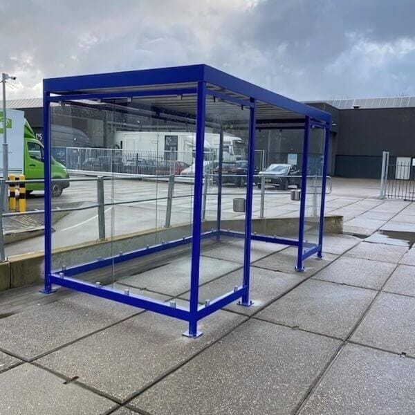 Rokershok kopen van 4x2 meter Rookabri met helder glas en blauw stalen frame op een nat bedrijventerrein met geparkeerde voertuigen.