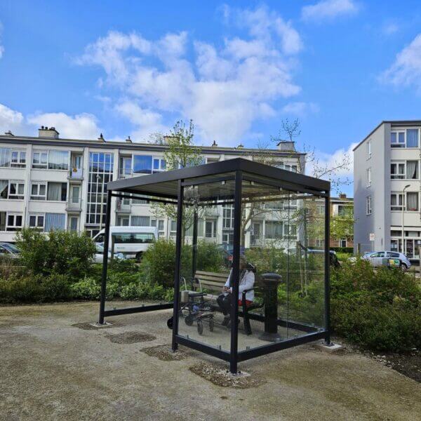 rokershok voor zorginstelling aanschaffen Rookoverkapping van glas met zitbank en asbak, geplaatst in een tuin bij een woonzorglocatie met appartementen.