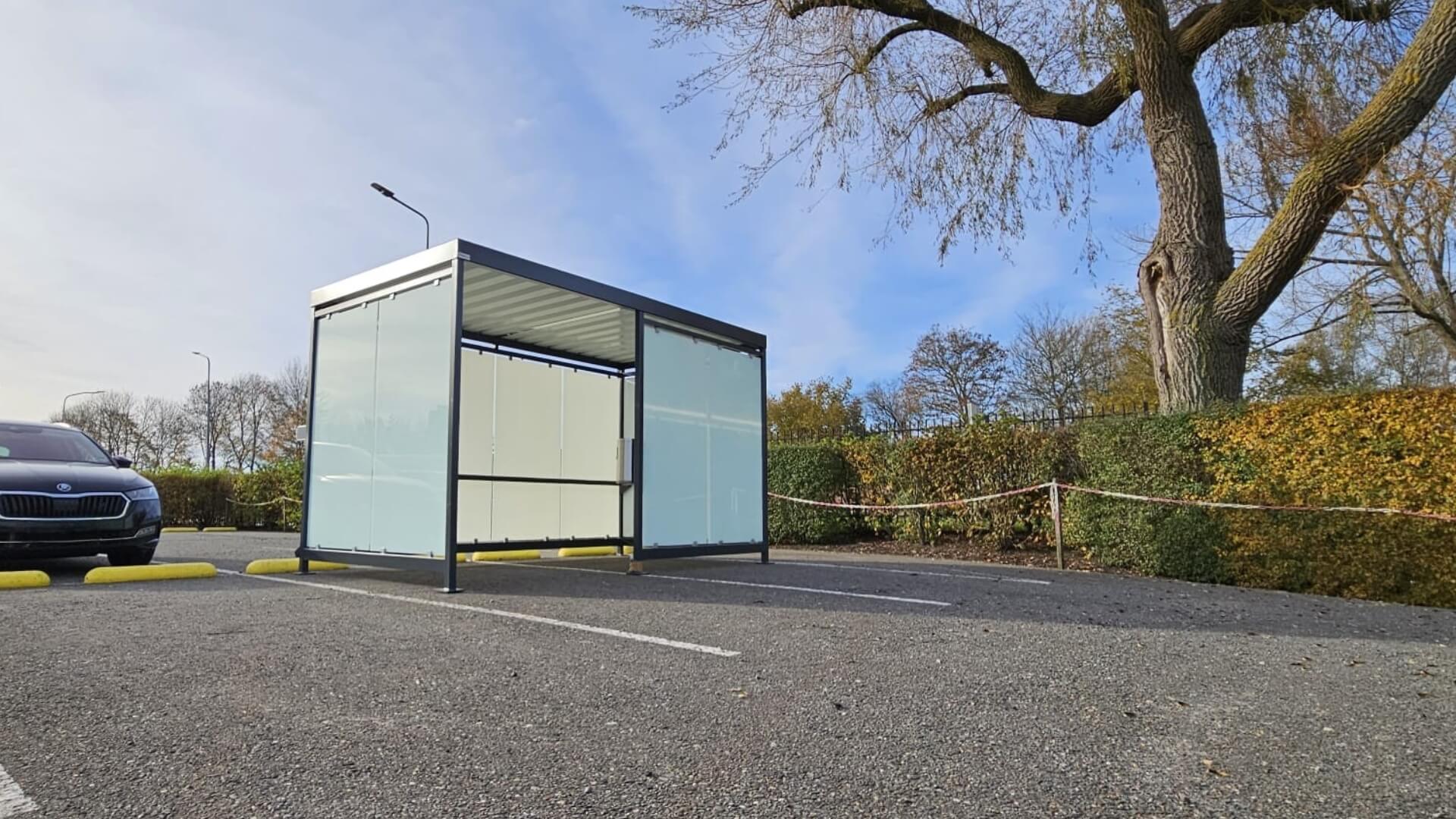 Rokersoverkapping Keulen van Umfeldt met stalen frame en matglazen wanden, geplaatst op parkeerterrein in Bedum, duurzame rookruimte voor buitengebruik, leverbaar in Nederland en België.