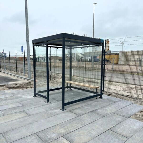rookabri aanschaffen Glazen rookabri van 2x2 meter met zitbank en wandasbak, geplaatst op een betegeld terrein met hekwerk op achtergrond.