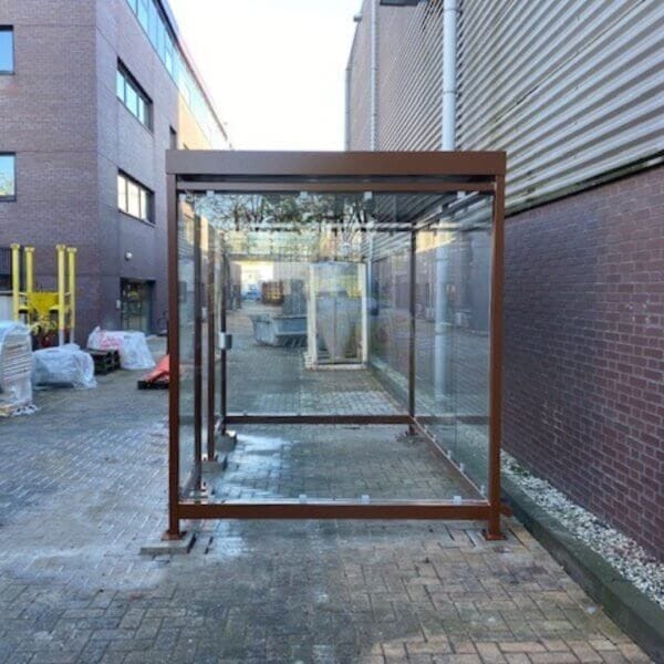 Rokersruimte met glazen wanden en een frame in cortenstaal-look, geplaatst op een verharde ondergrond tussen bedrijfsgebouwen.