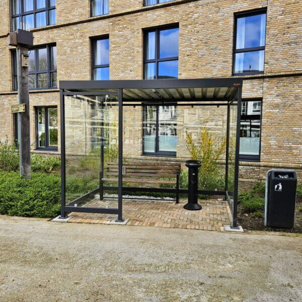 rookabri voor zorginstelling direct kopen Rookoverkapping van 3x2 meter met glazen wanden, bankje en asbak, geplaatst bij een gebouw met bakstenen gevel.
