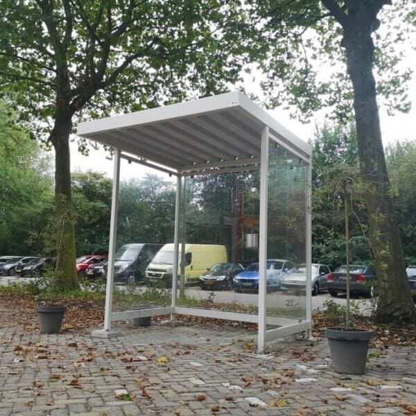 Witte rokershok van 2x2 meter met glazen wanden en metalen frame, geplaatst op een bestrate ondergrond en omringd door bomen.