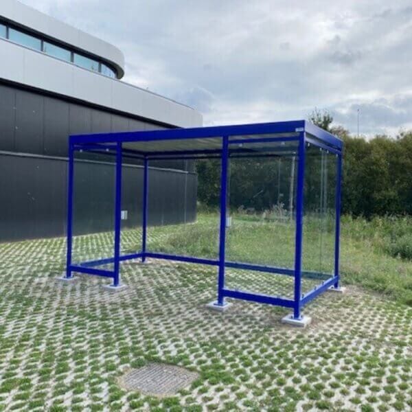 Functionele blauwe rookoverkapping voor buiten kopen Blauwe rookoverkapping met ESG glazen wanden, geplaatst op een groene open ruimte naast een modern gebouw.