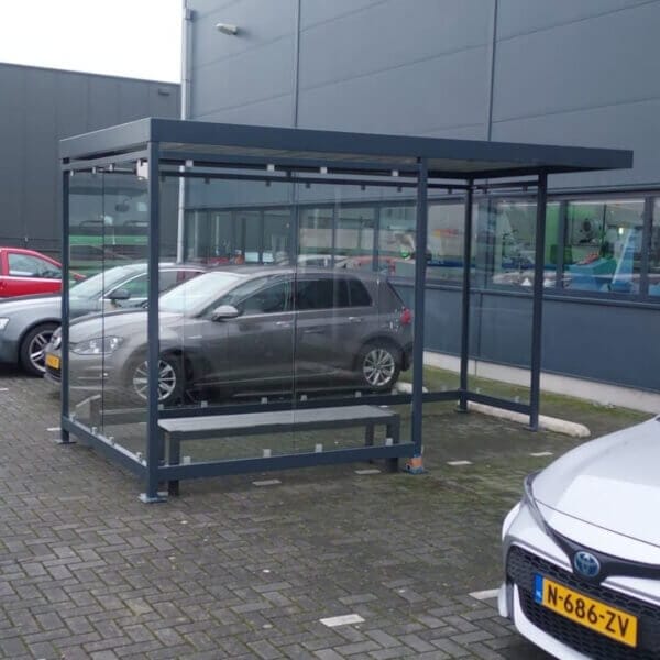 Prefab rookoverkapping online kopen Glazen rookoverkapping met ESG wanden en donkergrijs gepoedercoat stalen frame, voorzien van een geïntegreerde zitbank, geplaatst op een parkeerplaats bij een bedrijfsgebouw.