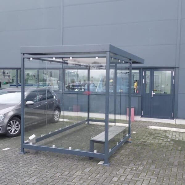 Prefab rookoverkapping aanschaffen Compacte rookoverkapping met ESG glazen wanden en een donkergrijs gepoedercoat stalen frame, voorzien van een eenvoudige zitbank, geplaatst op een parkeerplaats bij een bedrijfsgebouw.