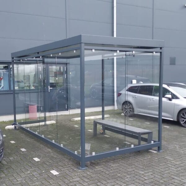 Prefab rookoverkapping kopen Rookoverkapping met ESG glazen wanden en een donkergrijs gepoedercoat stalen frame, geplaatst op een parkeerterrein en voorzien van een metalen zitbank.
