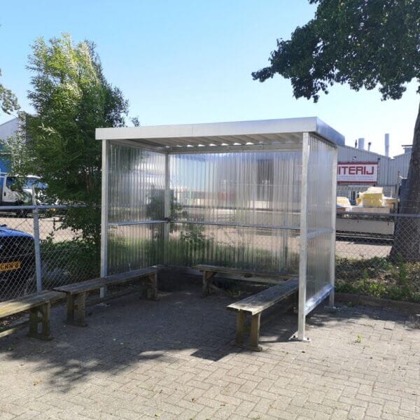 Bouwpakket rookoverkapping in verzinkte uitvoering met polycarbonaat zij- en achterwand, geplaatst op een bestrate ondergrond met houten banken