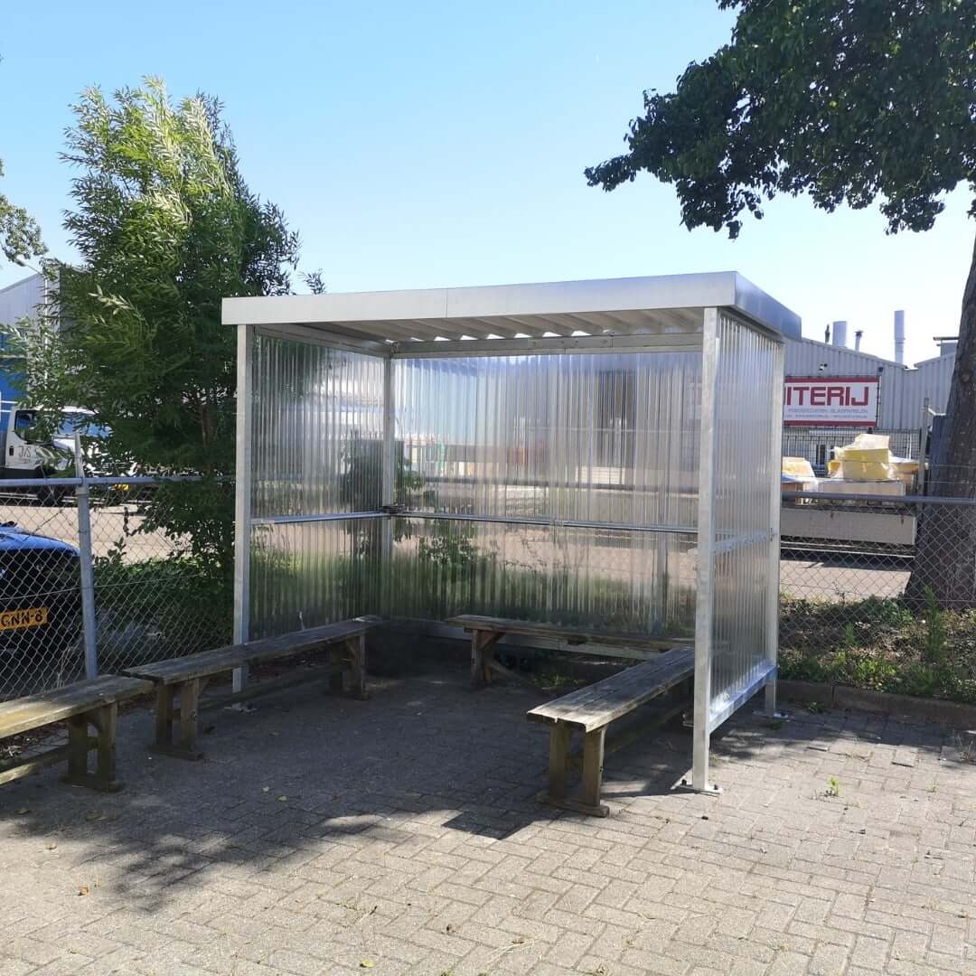 Bouwpakket rookoverkapping in verzinkte uitvoering met polycarbonaat zij- en achterwand, geplaatst op een bestrate ondergrond met houten banken