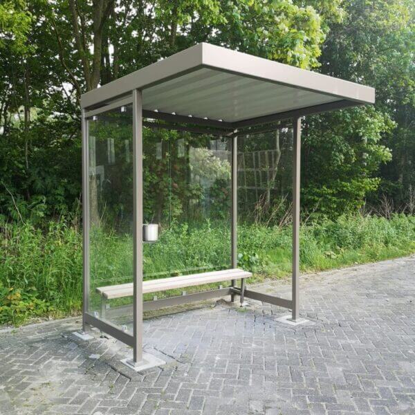 Compacte rookoverkapping met glazen wanden en een stevig metalen frame in een chique bruin/grijze kleur, voorzien van een houten zitbank en wandasbak. Geplaatst op een betegelde ondergrond naast een groene omgeving.