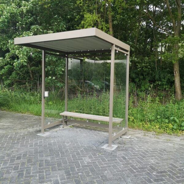 Buitengeplaatste rookoverkapping in een elegante bruin/grijze kleur, voorzien van ESG-glazen wanden, een comfortabele houten zitbank en een praktische wandasbak. Geplaatst op een betegelde ondergrond naast een groene, natuurlijke omgeving.