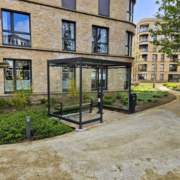 rookabri-keulen-3x2m-direct-online-bestellen Glazen rookabri met bankje en asbak, geplaatst in een verzorgde binnentuin bij een moderne zorginstelling.