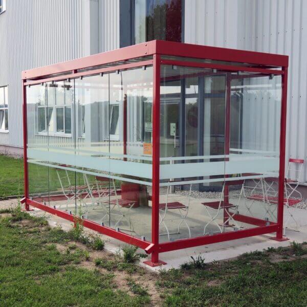 Rookhok kleur rood voor buiten kopen Rode rookhok voor buiten met ESG glazen wanden voorzien van zeefdruk, inclusief verzinkt en gepoedercoat stalen frame, geplaatst naast een bedrijfsgebouw.