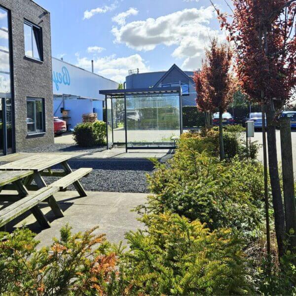 rookhok-kopen-voor-buiten Rookahok met glazen wanden en stalen frame op een bedrijventerrein tussen groen en zitbanken.