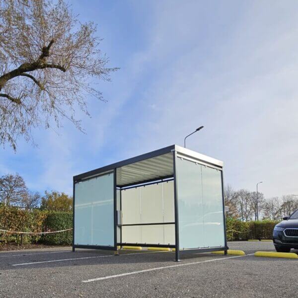 Rookoverkapping van Umfeldt aanschaffen Vrijstaande rookoverkapping van glas en metaal op een parkeerplaats, met zicht op een zwarte auto en lantaarnpaal.