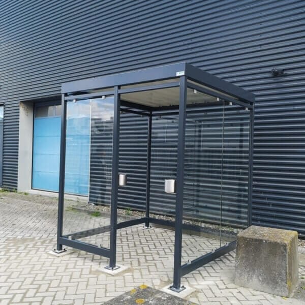 Donkere rookabri met glazen wanden en wandasbakken online aanschaffen Donkere rookabri geplaatst op bestrating, voorzien van twee wandasbakken en een transparante glazen ombouw.