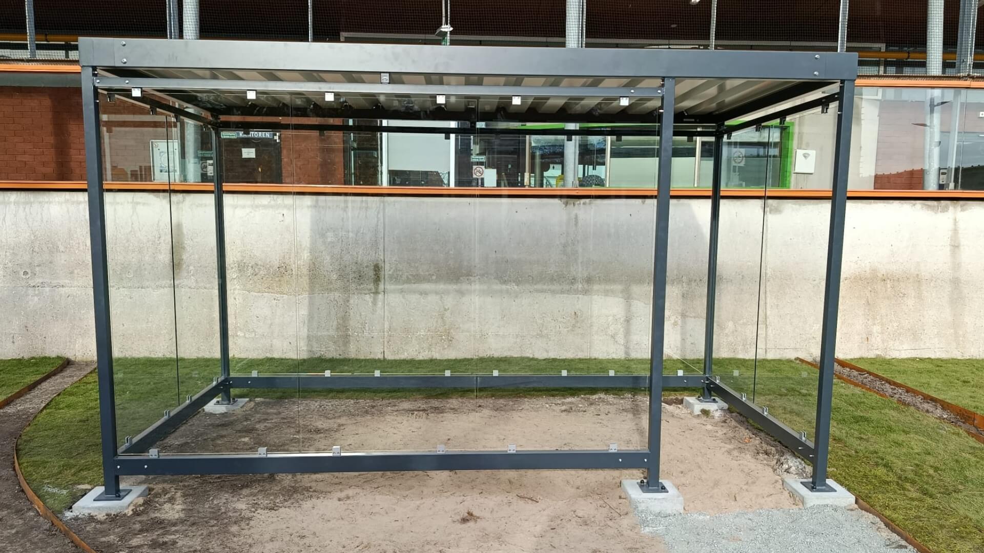 Vooraanzicht van een rookoverkapping van Umfeldt, met een frame van verzinkt staal en wanden van ESG glas. De overkapping staat op een zanderige met grind bedekte ondergrond tegen een betonnen wand bij COA Den Helder.