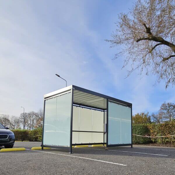 Rookruimte buiten van Umfeldt aanschaffen Vrijstaande rookruimte buiten met glazen wanden en metalen dak, geplaatst op een parkeerplaats naast een zwarte auto.