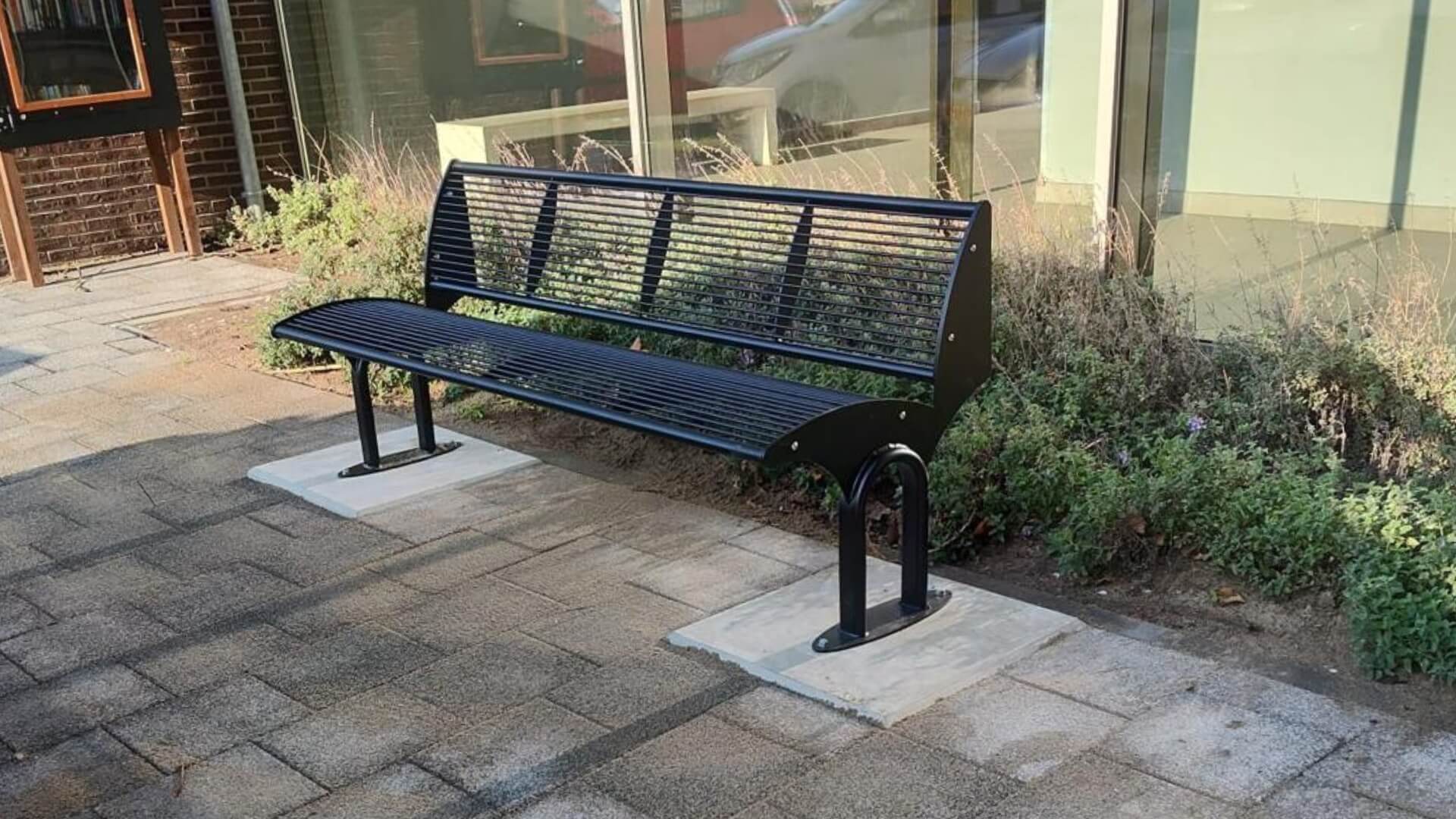 Zwarte stalen buitenbank Carinzia met horizontale lamellen, verankerd op betonnen platen naast een moderne glasgevel.