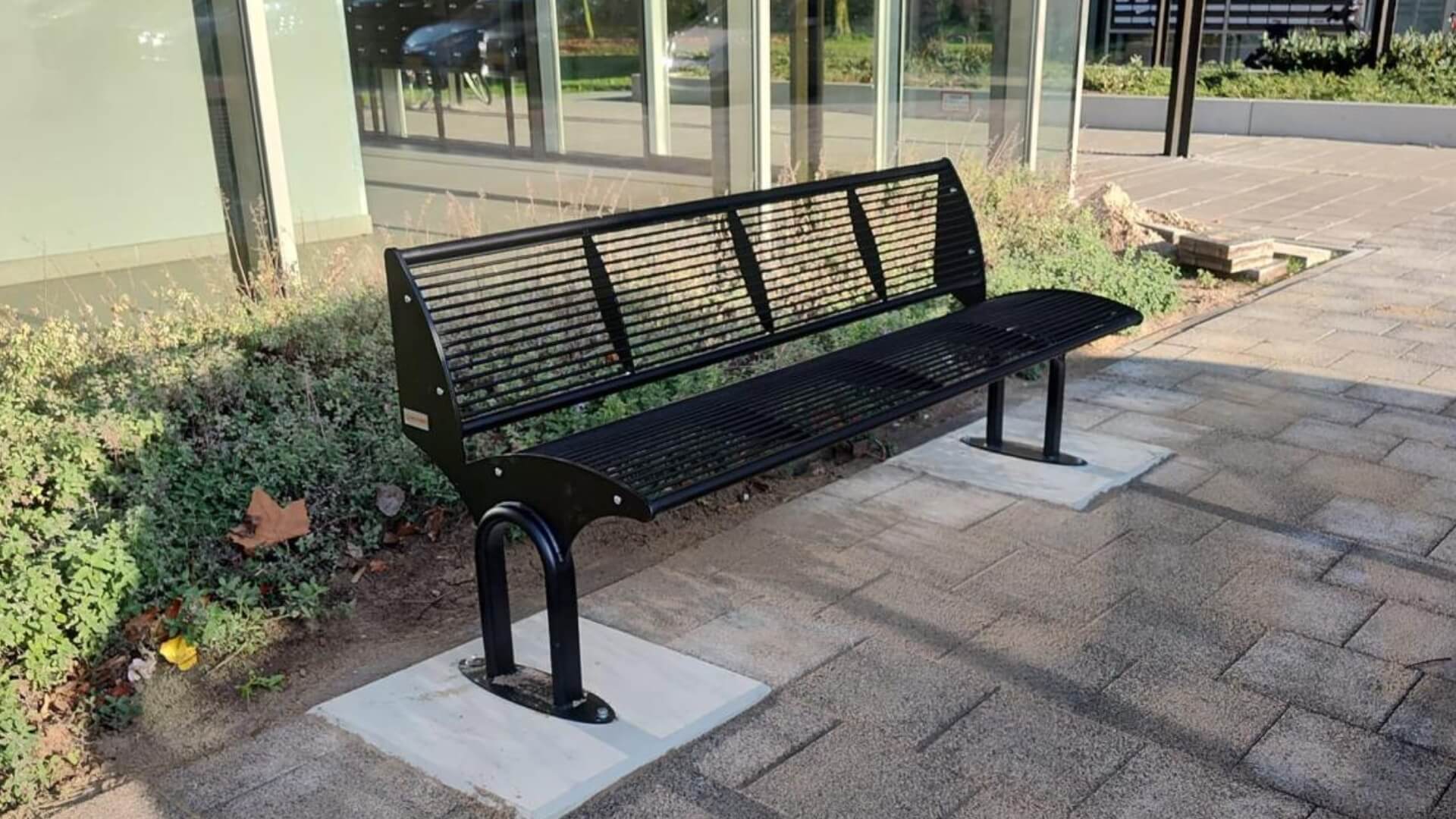 Zwarte stalen buitenbank Carinzia met horizontale lamellen, stevig verankerd op betonnen platen naast een moderne glasgevel.