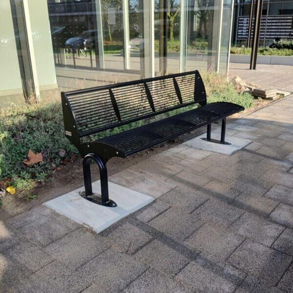 Stalen buitenbank bestellen voor openbare ruimtes Zwarte stalen buitenbank Carinzia met poedercoating, verankerd op betonnen platen naast een gebouw met glasgevel.