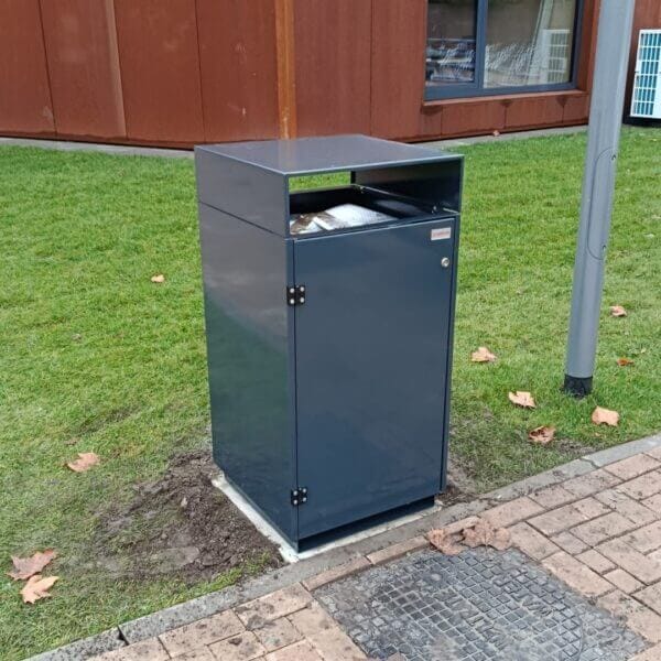 Afvalbak buiten direct aanschaffen voor schoolpleinen Donkergrijze afvalbak van verzinkt staal met boveninworp, geplaatst langs een voetpad op het grasveld bij Reggesteyn in Nijverdal. De afvalbak is uitgevoerd in kleur RAL 7016.