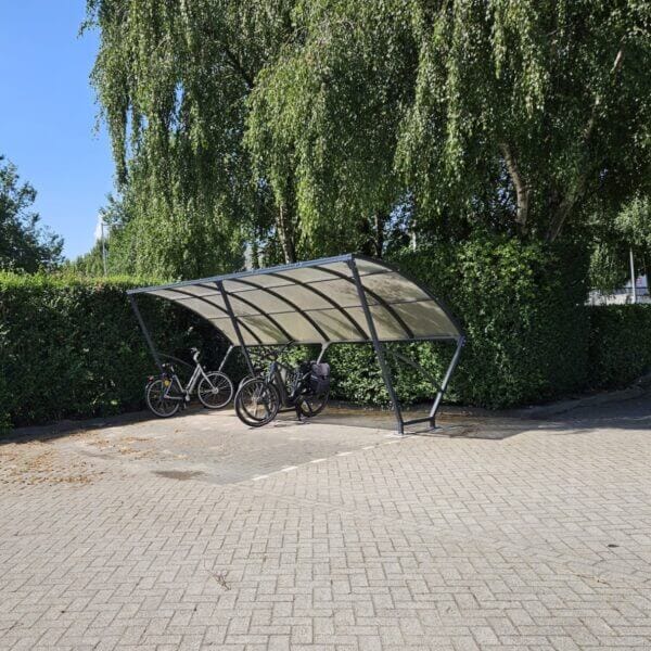Boogkap fietoverkapping van 5 meter aanschaffen Fietsoverkapping met gebogen dak en zwart stalen frame, geplaatst op een betegeld terrein tegen een groene haag. Onder de overkapping staan drie fietsen geparkeerd in de schaduw van grote bomen.