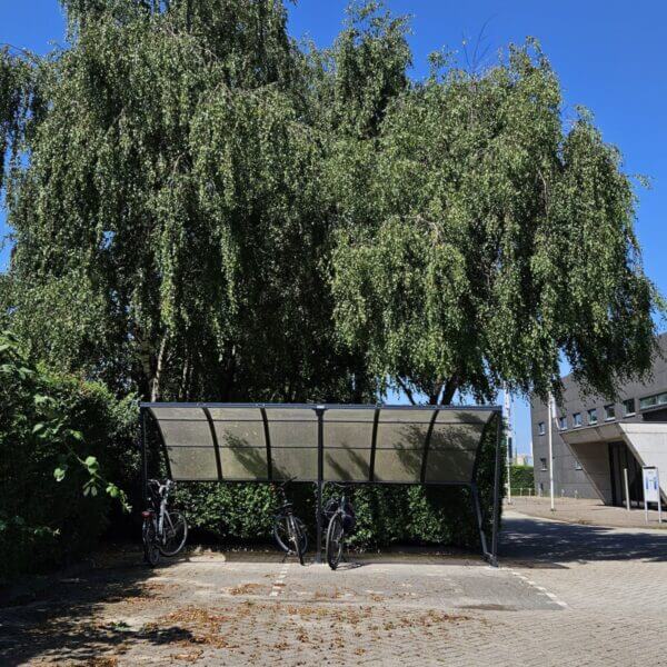 Fietsoverkapping 5 meter direct online bestellen Fietsenstalling met gebogen transparant dak en zwart metalen frame, geplaatst op een betegeld terrein tussen bomen en struiken. Drie fietsen staan onder de overkapping, met een kantoorgebouw op de achtergrond.