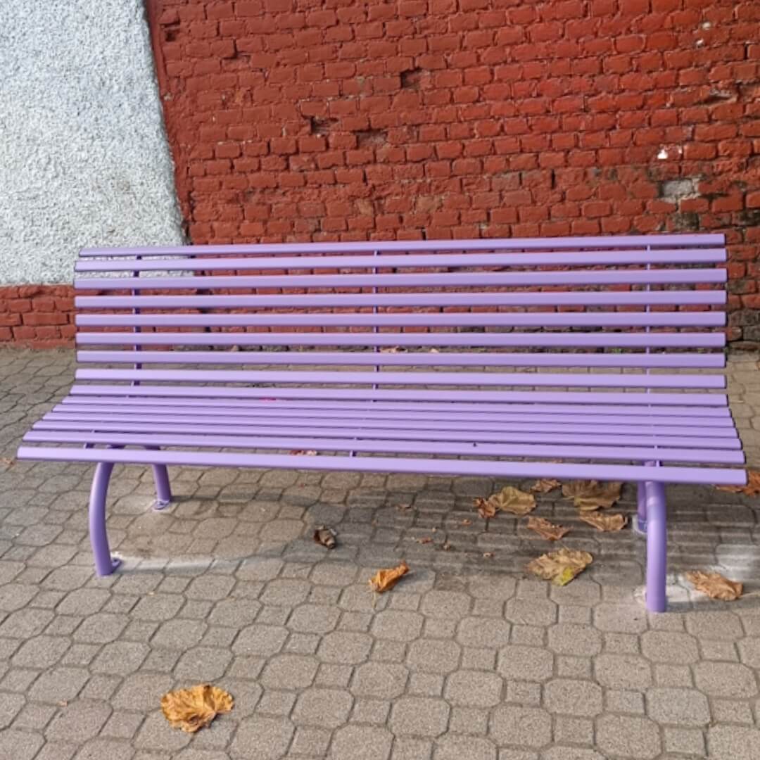 Lila kleur buitenbank van staal staat op een bestrate stoep, met op de achtergrond een muur van rode bakstenen.