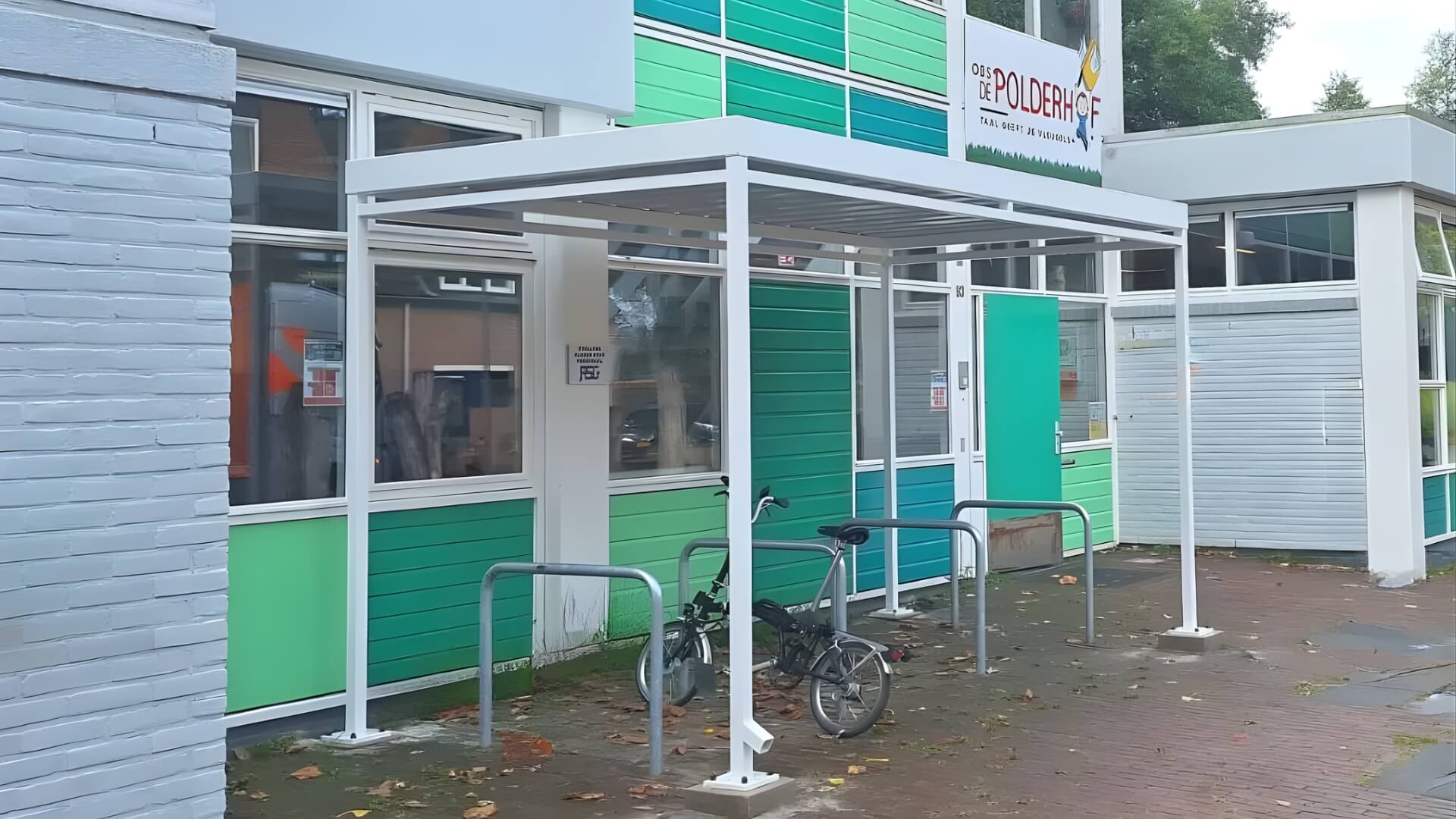 Witte fietsoverkapping bij basisschool De Polderhof in Almere, voorzien van fietsenrekken en geplaatst naast de ingang van het schoolgebouw met groene gevelpanelen.