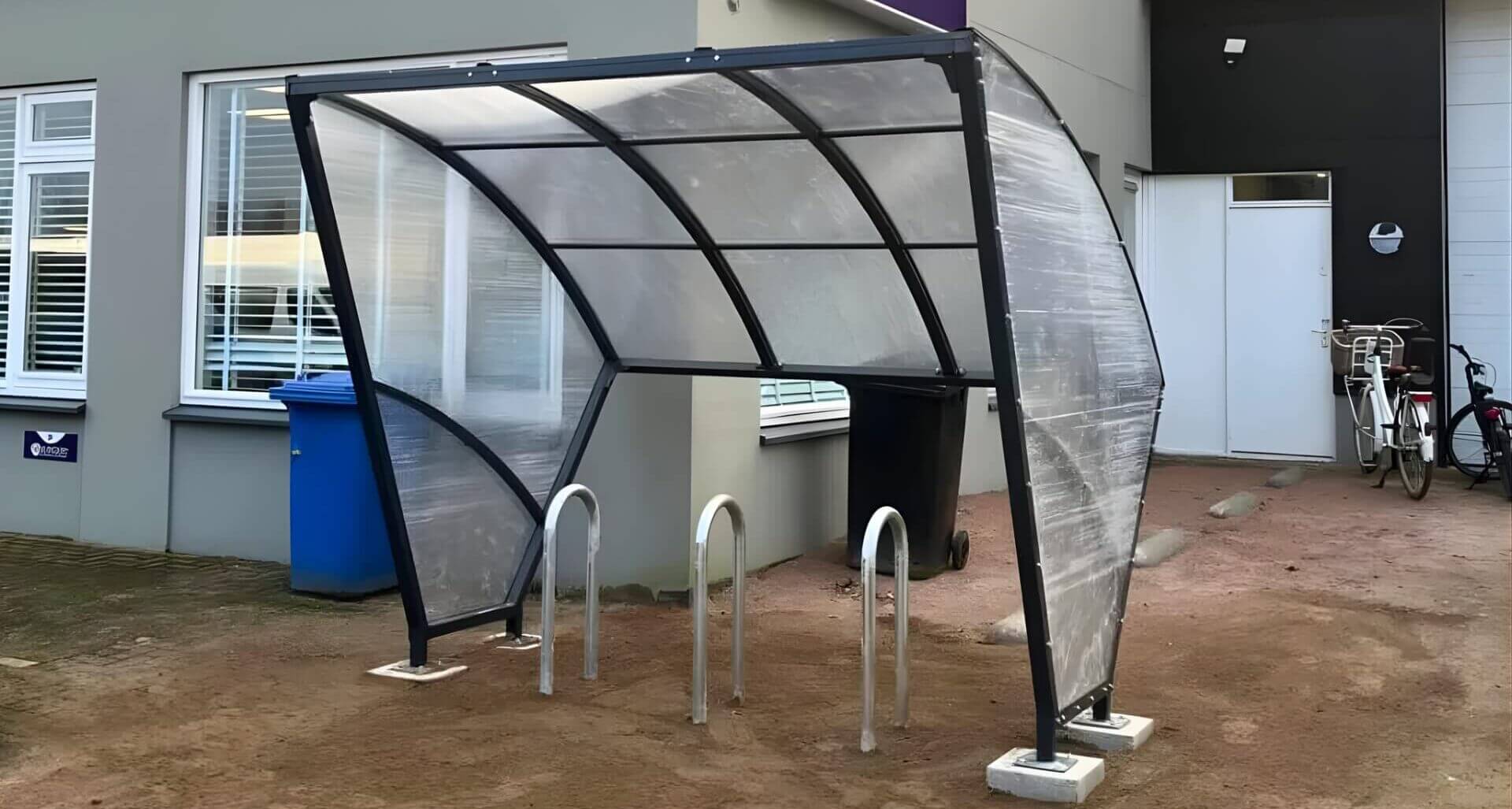 Compacte fietsoverkapping met transparant gebogen dak en zwart stalen frame, geplaatst naast een modern bedrijfsgebouw met fietsenrekken eronder.