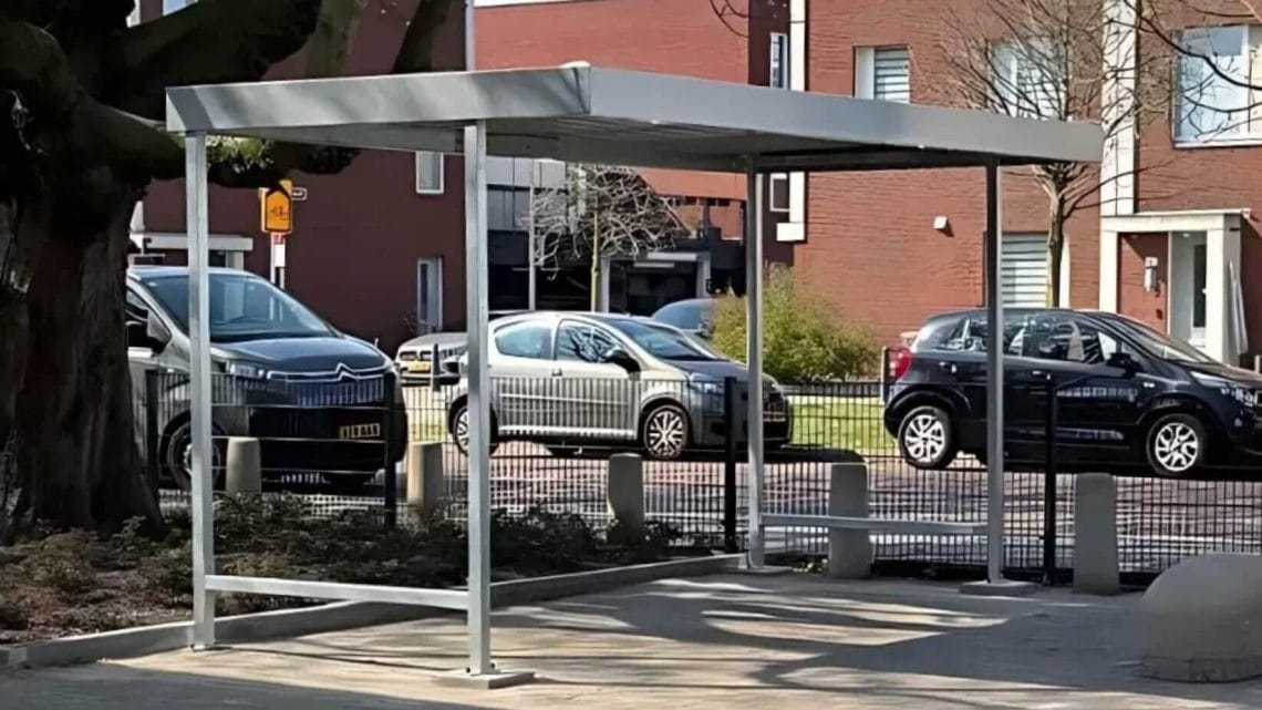 Moderne metalen fietsoverkapping op een betegeld plein naast een boom, geplaatst bij een wooncomplex met geparkeerde auto’s op de achtergrond.