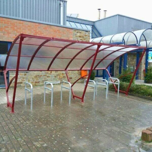 Rode fietsenoverkapping van 5 meter. Fietsoverkapping rood 5 meter voorzien van fietsenrekken, geplaatst op tegels bij een instelling.