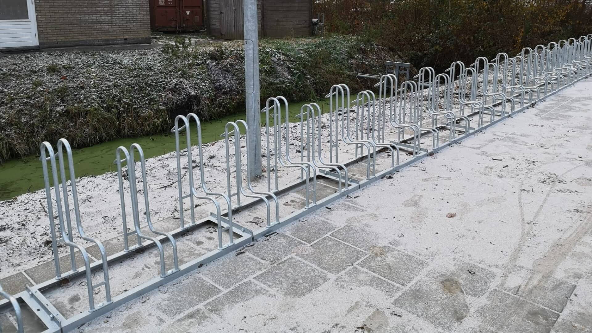 Rij fietsrekken van verzinkt staal op een besneeuwd betegeld terrein naast een sloot, geplaatst bij HS88 in Hoogezand. De rekken bieden plek aan meerdere fietsen en zijn stevig verankerd in de grond.