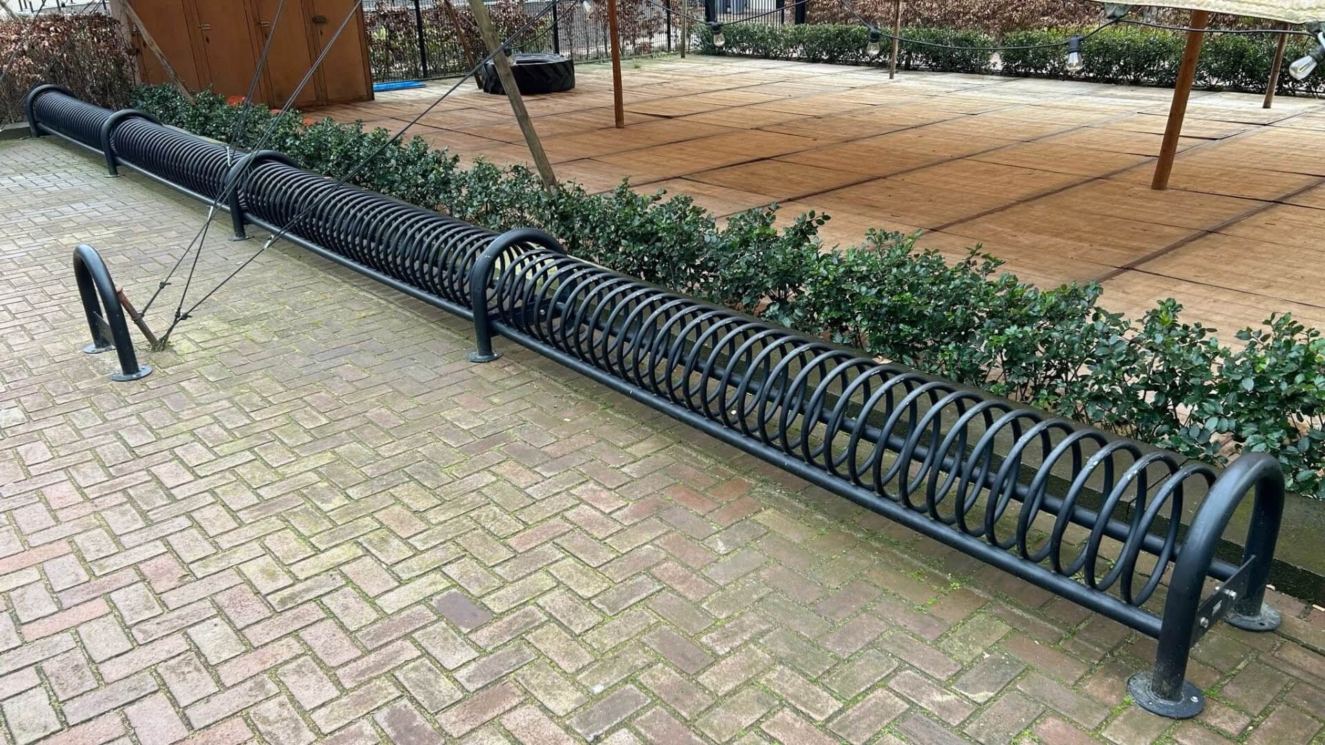Lang, zwart stalen fietsenrek met spiraalvormig ontwerp, geschikt voor meerdere fietsen, geplaatst op een betegelde ondergrond naast een haag. 