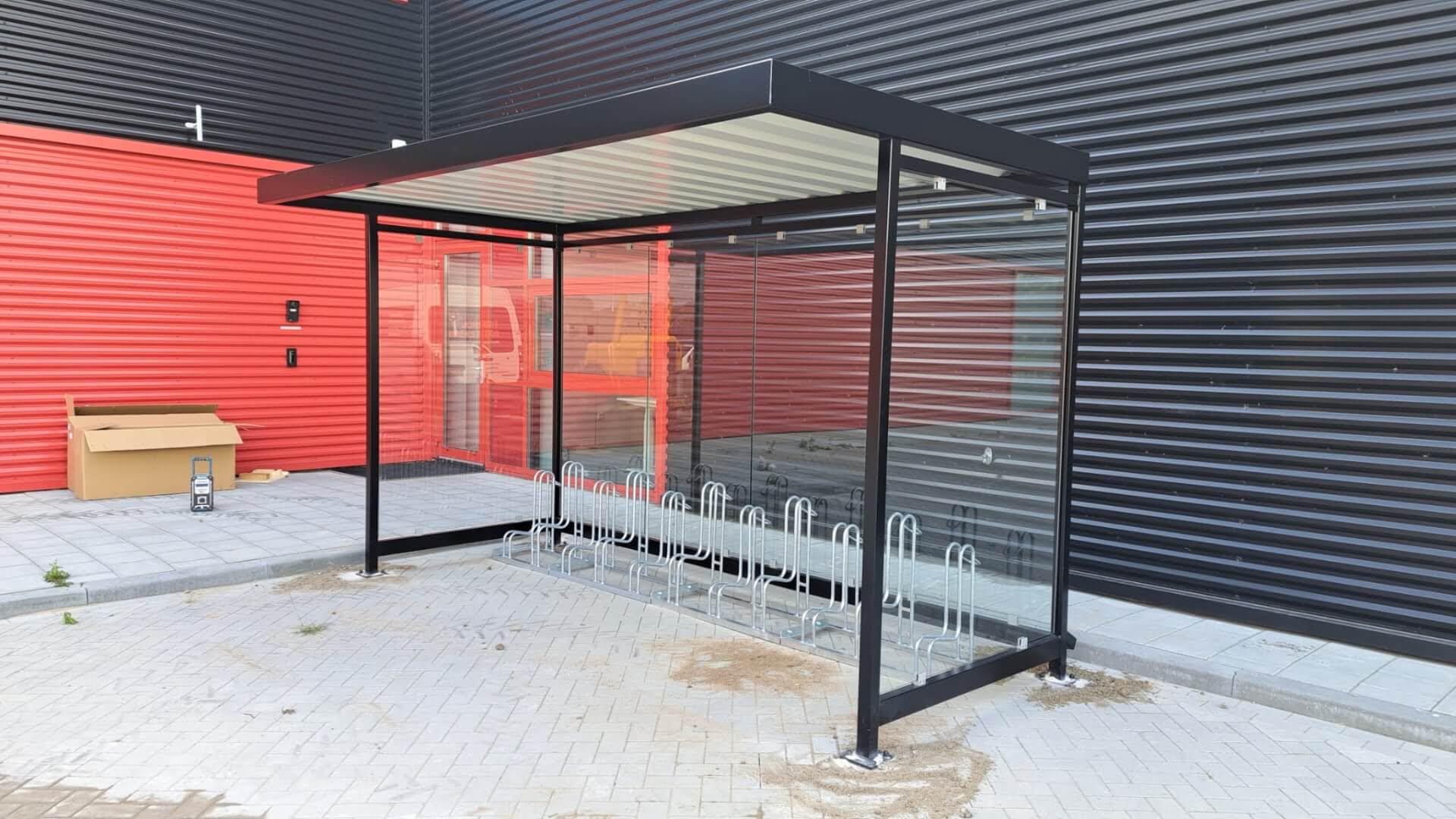 Fietsoverkapping met zwart metalen frame en transparante zijwanden, geplaatst tegen een zwart-rood gebouw. Onder de overkapping staat een rij zilverkleurige fietsenrekken geschikt voor het stallen van meerdere fietsen.